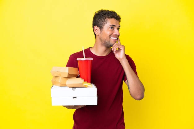 Młody brazylijski mężczyzna trzymający pizze i hamburgery na białym tle, myślący o pomyśle, patrząc w górę