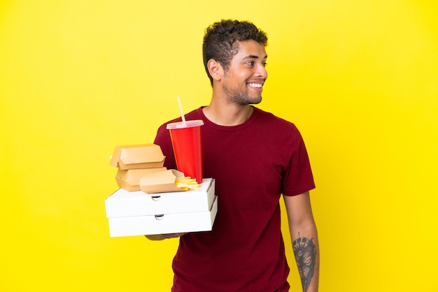 Młody brazylijski mężczyzna trzymający pizze i hamburgery na białym tle, patrząc w bok i uśmiechnięty
