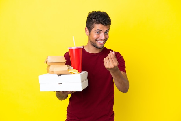 Młody brazylijski mężczyzna trzymający pizze i hamburgery na białym tle robiący gest pieniędzy