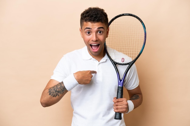 Młody brazylijski przystojny mężczyzna grający w tenisa na białym tle na beżowym tle z niespodzianką wyrazem twarzy