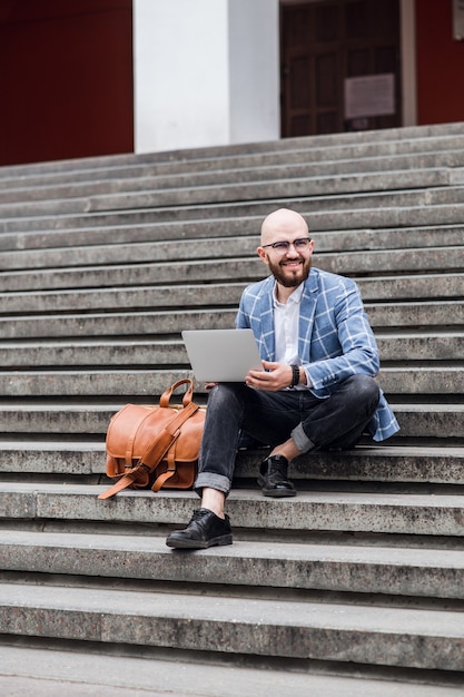 Młody brodaty biznesmen siedzi na schodach za pomocą laptopa i patrzy na jego screenhipster mężczyzna pracuje na czacie online, sprawdzając pocztę e-mail