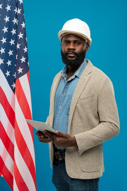 Młody, brodaty inżynier afroamerykański używający cyfrowego tabletu, stojący na tle flagi z gwiazdami i paskami oraz niebieskiej flagi