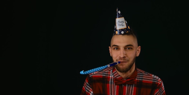Młody brodaty kaukaski mężczyzna nosi świąteczny kapelusz z napisem happy party i dmucha w papierową fajkę