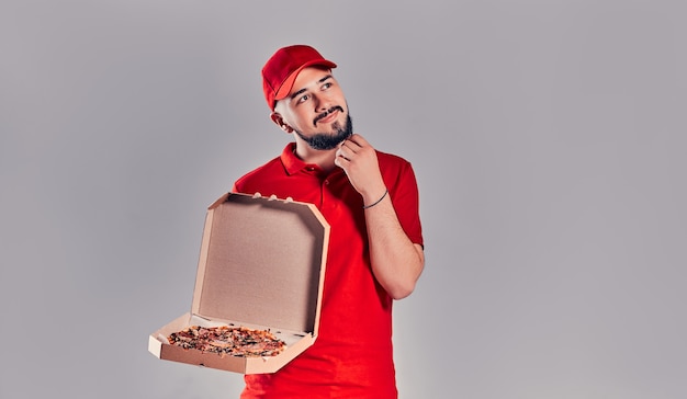 Zdjęcie młody brodaty mężczyzna dostawy w czerwonym mundurze myśli o czymś, co trzyma pudełko po pizzy na białym tle na szarym tle.
