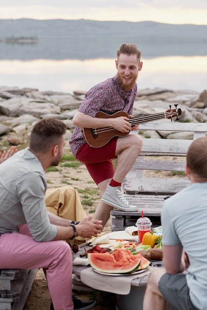 Młody brodaty mężczyzna gra na gitarze i uśmiecha się radośnie spędzając letni wieczór z przyjaciółmi na świeżym powietrzu nad jeziorem