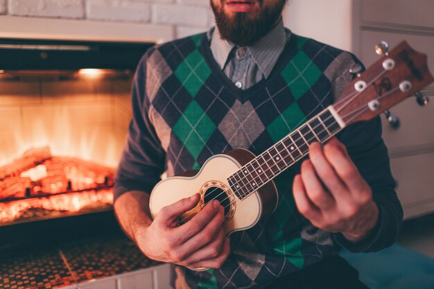 młody brodaty mężczyzna gra na ukulele w pokoju przy kominku