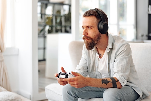 Młody brodaty mężczyzna grający w gry wideo siedząc na kanapie w domu
