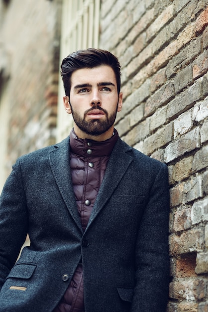 Młody brodaty mężczyzna jest ubranym brytyjskiego eleganckiego kostium w ulicie w miastowym tle.