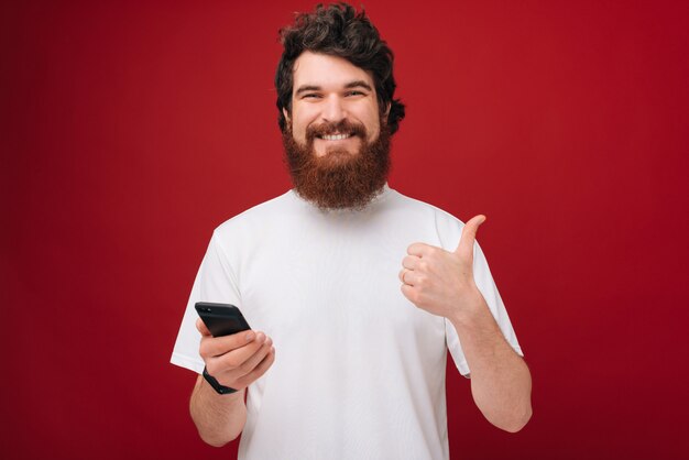 Młody brodaty mężczyzna nad czerwieni ściany mienia smartphone szczęśliwym z dużym uśmiechem robi ok znakowi, kciuk up