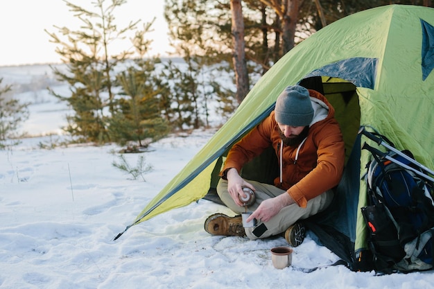 Młody brodaty mężczyzna odpoczywa w zimowych górach w pobliżu namiotu Podróżnik z brodą w czapce i ciepłej kurtce rozgrzewa się pijąc gorącą herbatę lub kawę po wędrówce Podróżniczy styl życia