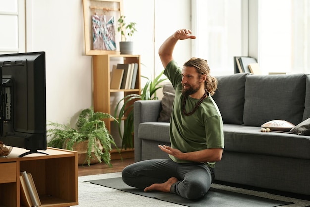 Młody brodaty mężczyzna ogląda program telewizyjny i robi ćwiczenia jogi w salonie w domu