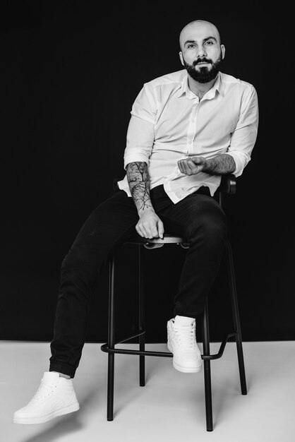 Młody brodaty mężczyzna w białej koszuli siedzi na czarnej powierzchni