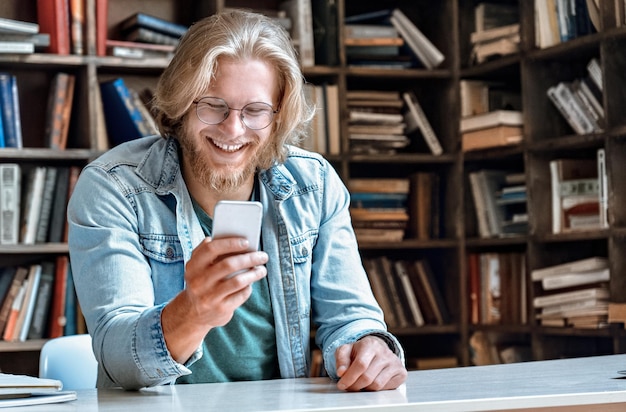 Młody brodaty mężczyzna w okularach na biurku bibliotecznym trzyma nowoczesny smartfon i śmieje się z ekranu