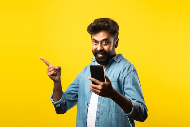 Młody Brodaty Mężczyzna Z Indii Używający Smartfona, Uśmiechający Się Podczas Rozmowy Lub Rozmowy Z Przyjacielem, Stojący Na żółto