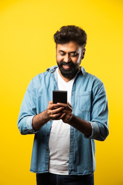 Młody brodaty mężczyzna z Indii używający smartfona, uśmiechający się podczas rozmowy lub rozmowy z przyjacielem, stojący na żółto