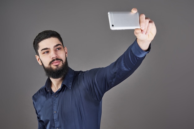 Młody brodaty modnisia biznesowy mężczyzna opowiada selfie fotografię z mądrze telefonem uśmiechniętym i patrzeje telefon na szarość