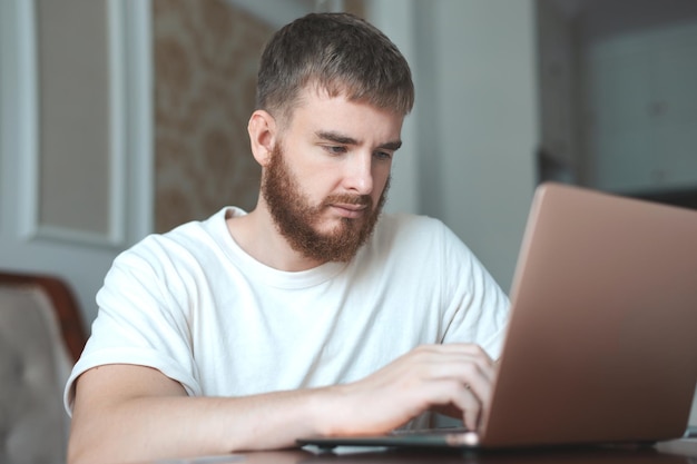 Młody brodaty skoncentrowany mężczyzna używa laptopa w domu w salonie z poważnym pisaniem na twarzy