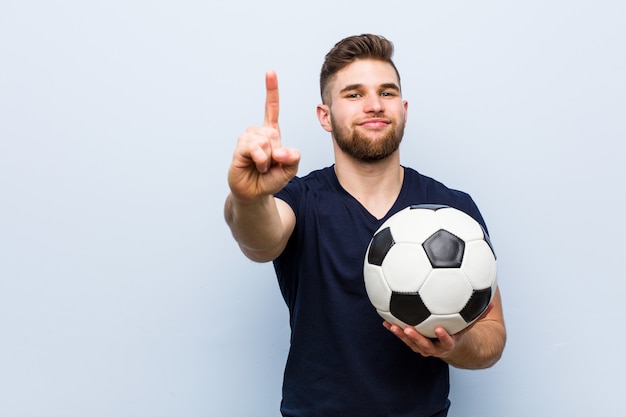 Młody caucasian mężczyzna trzyma piłki nożnej piłkę pokazuje liczbę jeden z palcem.