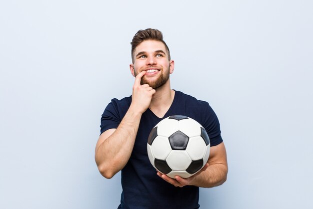 Młody caucasian mężczyzna trzyma piłki nożnej piłkę relaksował myśleć o coś patrzeje odbitkową przestrzeń.