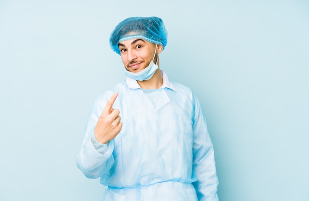 Młody chirurg arabski mężczyzna na białym tle na niebieskiej ścianie wskazując palcem na ciebie, jakby zapraszając, podejdź bliżej.