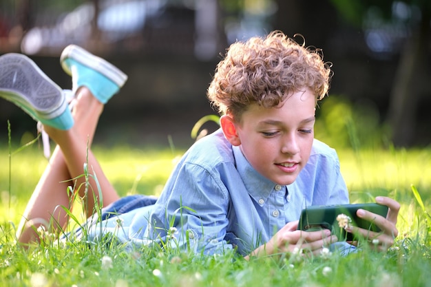 Młody chłopak gra na swoim smartfonie na świeżym powietrzu w letnim parku Uzależnienie od koncepcji gadżetów elektronicznych