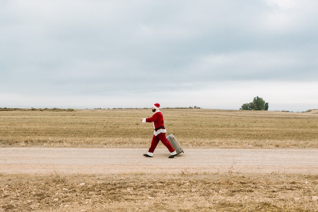 Młody chłopak przebrany za Świętego Mikołaja z walizką spacerujący po wiejskiej okolicy