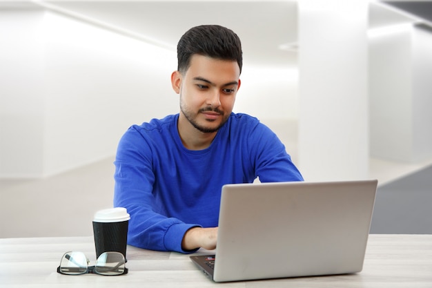 Młody chłopak siedzący przy laptopie w poszukiwaniu pracy w Internecie, robiący interesy w globalnej sieci przy filiżance kawy. W świetle