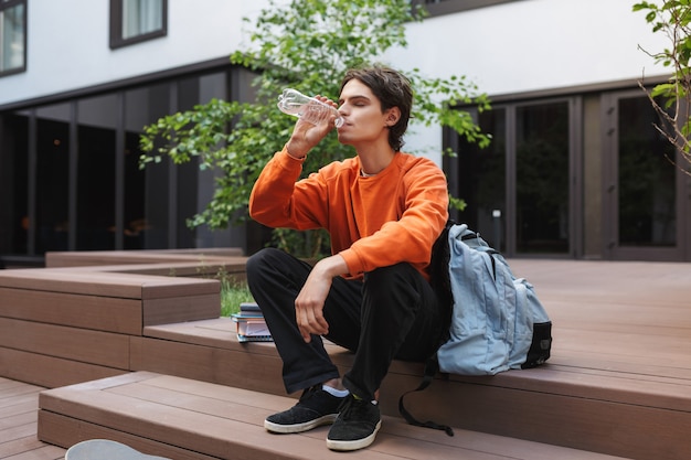 Młody chłopak siedzi i pije wodę z dużym plecakiem w pobliżu dziedzińca uniwersytetu