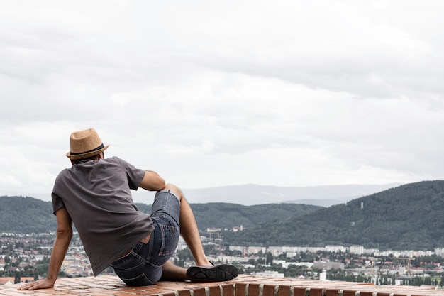 Młody chłopak siedzi na skraju wysokiego budynku i rozkoszuje się widokiem na góry