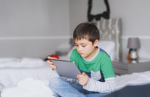 Młody chłopak używa tabletu, grając w gry internetowe z przyjacielem. Nauka w domu. Dziecko odrabia zadanie domowe online za pomocą cyfrowego pada w domu. Dziecko siedzi na łóżku i relaksuje się, oglądając kreskówki lub rozmawiając online z przyjacielem.