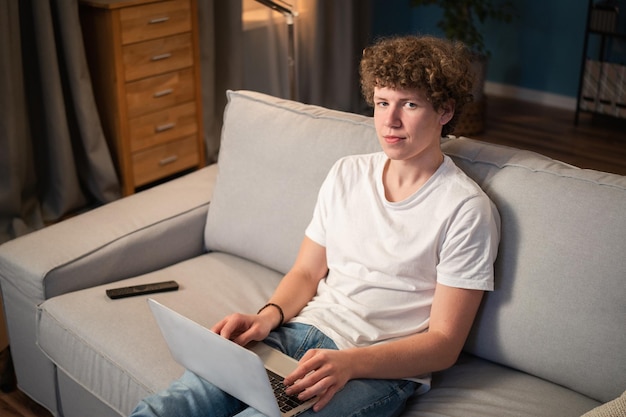 Młody chłopak z kręconymi włosami siedzi na kanapie w salonie ze swoim laptopem