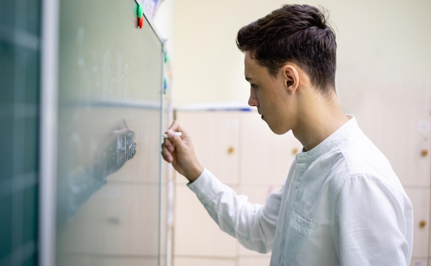 Młody ciemnowłosy student rasy kaukaskiej stoi przy tablicy i pisze kredą