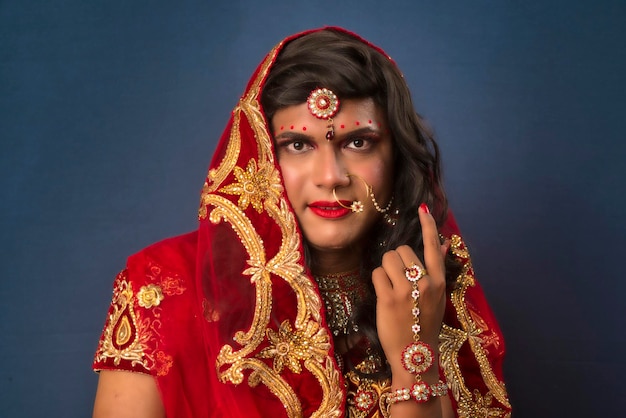 Młody crossdresser mężczyzna ubrany w indyjskie stroje ślubne panny młodej z makijażem ślubnym i pozowanie na szarym tle, patrząc modnie i efektownie