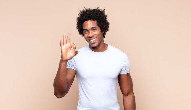 Młody czarny afro mężczyzna czuje się szczęśliwy, zdumiony, zadowolony i zaskoczony, pokazując gesty w porządku i kciuki w górę, uśmiechając się