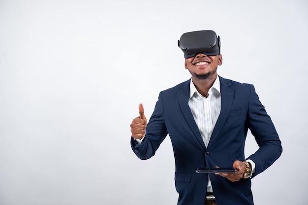 Młody czarny mężczyzna noszący zestaw słuchawkowy wirtualnej rzeczywistości i trzymający telefon uśmiecha się i pokazuje kciuk w górę