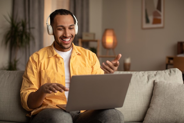 Młody czarny mężczyzna w słuchawkach za pomocą laptopa do czatu online podczas wideokonferencji lub biznesu