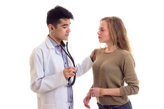Młody czarujący lekarz w białej sukni ze stetoskopem słuchający bicia serca młodej kobiety