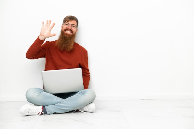 Młody czerwonawo-kaukaski mężczyzna z laptopem na białym tle pozdrawiający ręką ze szczęśliwym wyrazem twarzy