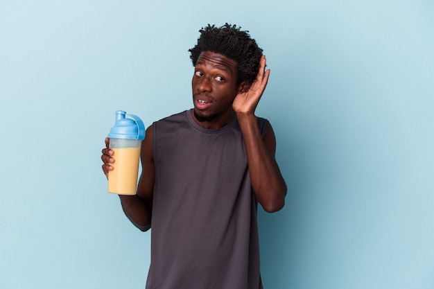 Młody człowiek african american gospodarstwa koktajl mleczny białka na białym tle na niebieskim tle próbuje słuchać plotek.