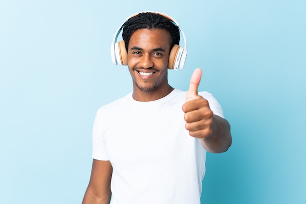 Młody człowiek African American z warkoczami na białym tle na niebieskim tle słuchania muzyki iz kciukiem do góry