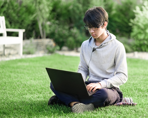 Młody człowiek azjatyckich w okularach działa na laptopie na trawniku na świeżym powietrzu