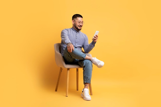 Młody Człowiek Azji Za Pomocą Smartfona Siedząc W Krześle Na żółtym Tle
