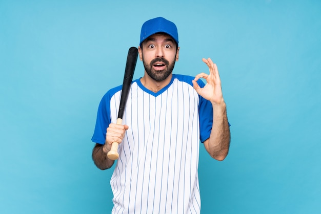 Młody człowiek bawić się baseballa nad odosobnioną błękit ścianą zaskakującą i pokazuje ok znaka