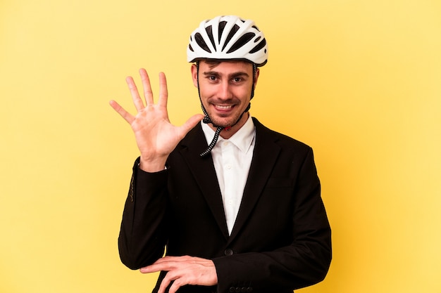 Młody człowiek biznesu kaukaski sobie kask rowerowy na białym tle na żółtym tle uśmiechający się wesoły pokazując numer pięć palcami.