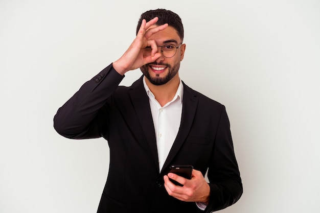 Młody człowiek biznesu rasy mieszanej posiadający telefon komórkowy mężczyzna na białym podekscytowany utrzymując ok gest na oko.