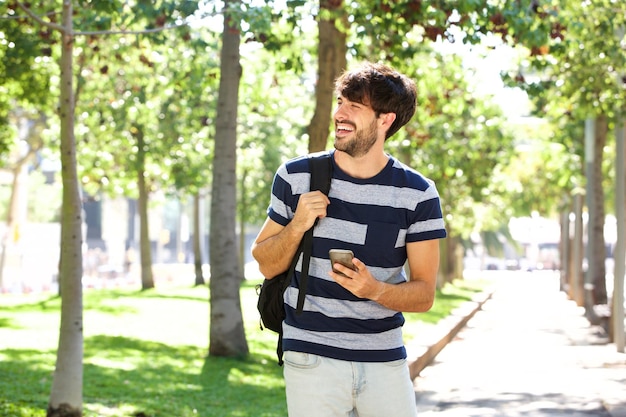 Młody człowiek chodzący z telefonem komórkowym i torbą w parku
