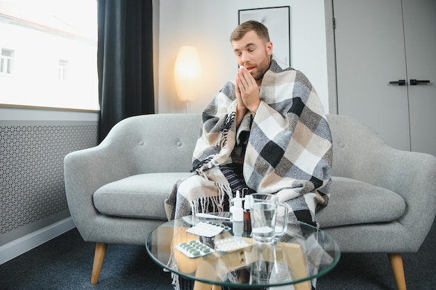 Zdjęcie młody człowiek cierpiący na przeziębienie i grypę lub alergię siedzi w domu owinięty w koc i wyciera nos chusteczkami, podczas gdy ma silny ból głowy koncepcja opieki zdrowotnej