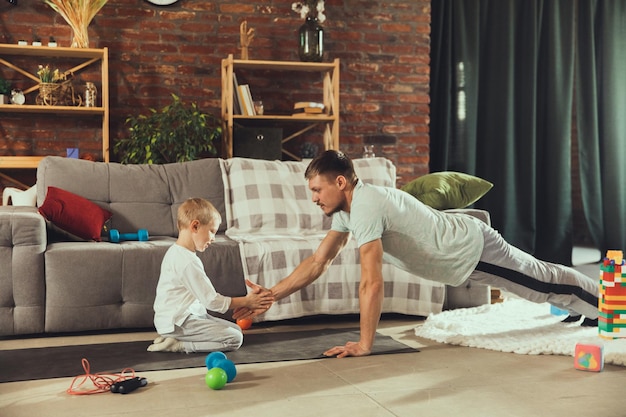 Młody człowiek ćwiczenia fitness, aerobik, joga w domu, sportowy styl życia. Aktywność, podczas gdy jego dziecko bawi się w tle, domowa siłownia.