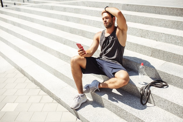 Młody człowiek ćwiczy na zewnątrz. Skoncentrowany sportowiec po treningu siada na schodach i odpoczywa. Trzymając telefon w dłoni i patrząc na niego.