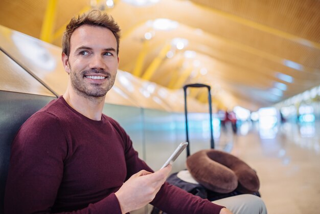 Młody człowiek czeka słuchanie muzyki i korzystanie z telefonu komórkowego na lotnisku z walizką.